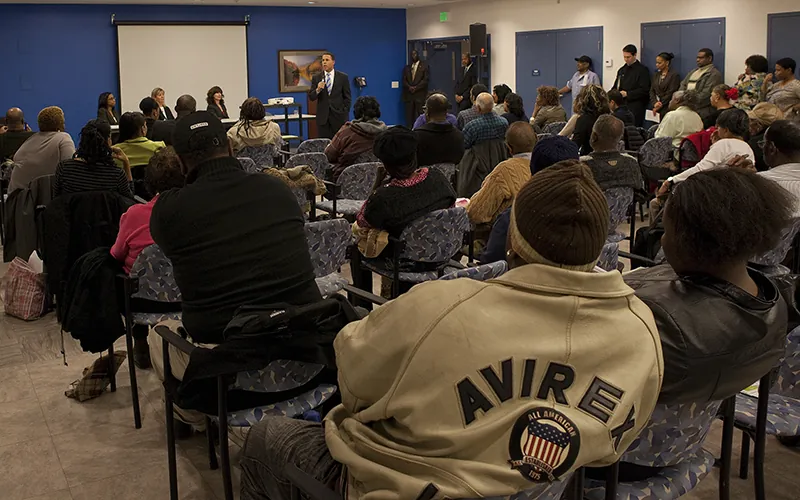 A large group of people sit in chairs and stand against the wall in a conference room. Three people wearing suits sit at a table at the front of the room, and a fourth person wearing a suit  stands beside them, addressing the crowd.