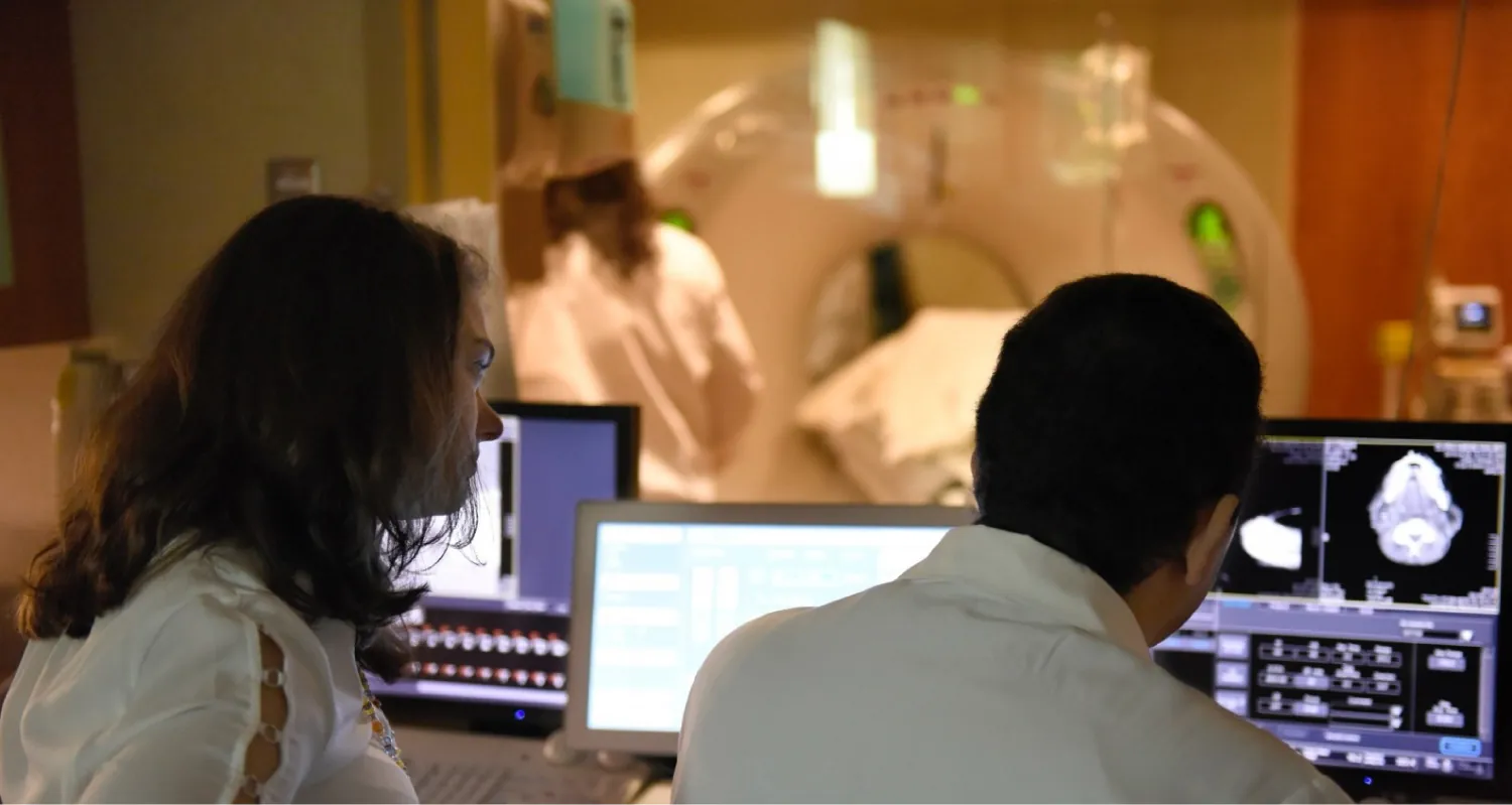 Two people looking at computers with brain images with an imaging equipment room in the background.