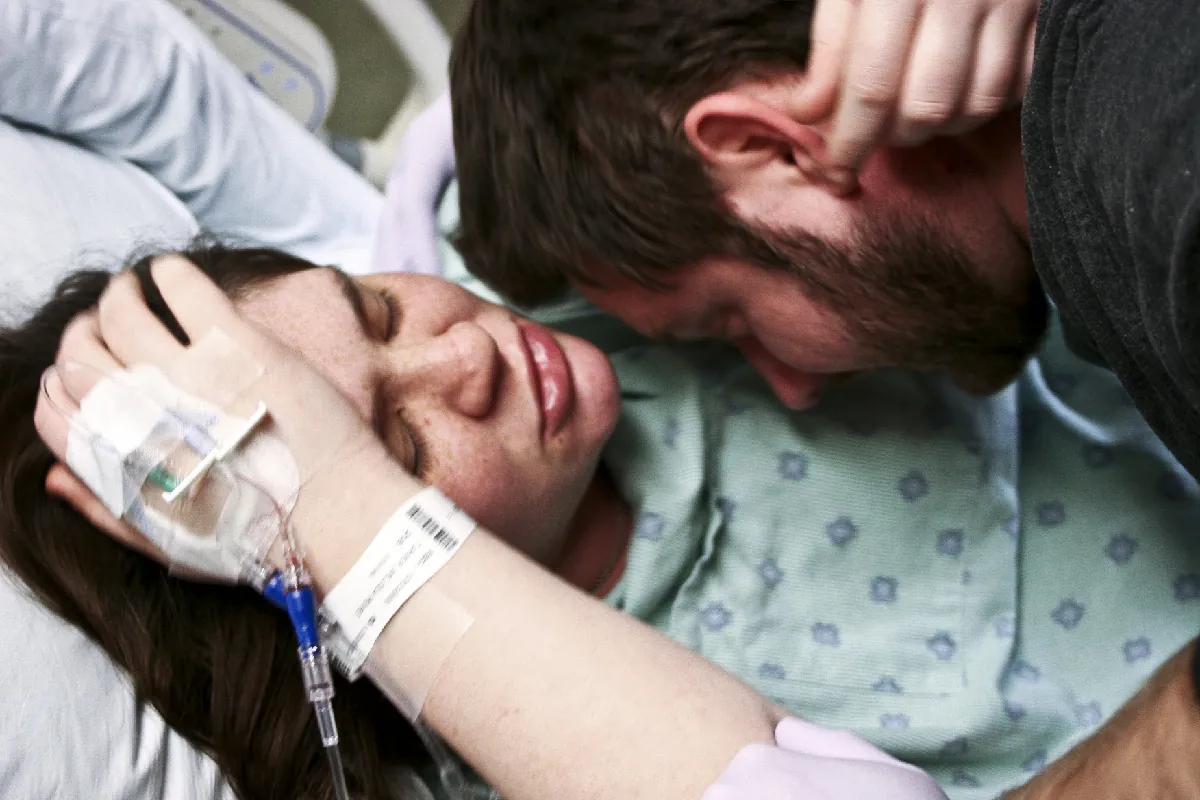 A color photograph of a patient wearing a hospital gown lying down in a hospital bed holding her head with her hands.
