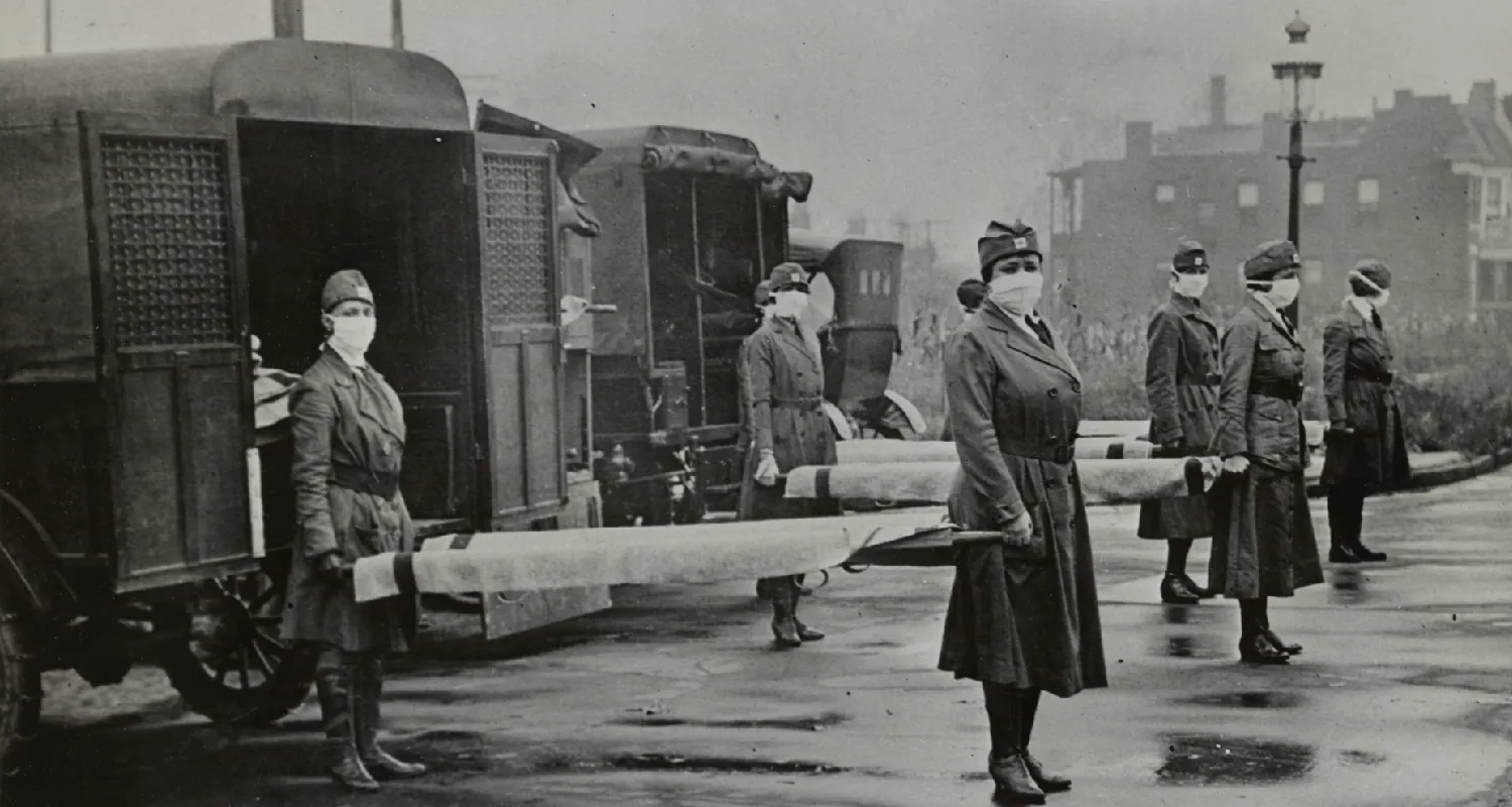 In a historical photo, nurses stand in pairs holding stretchers in front of ambulances. The nurses all wear paper face masks over their nose and mouth.