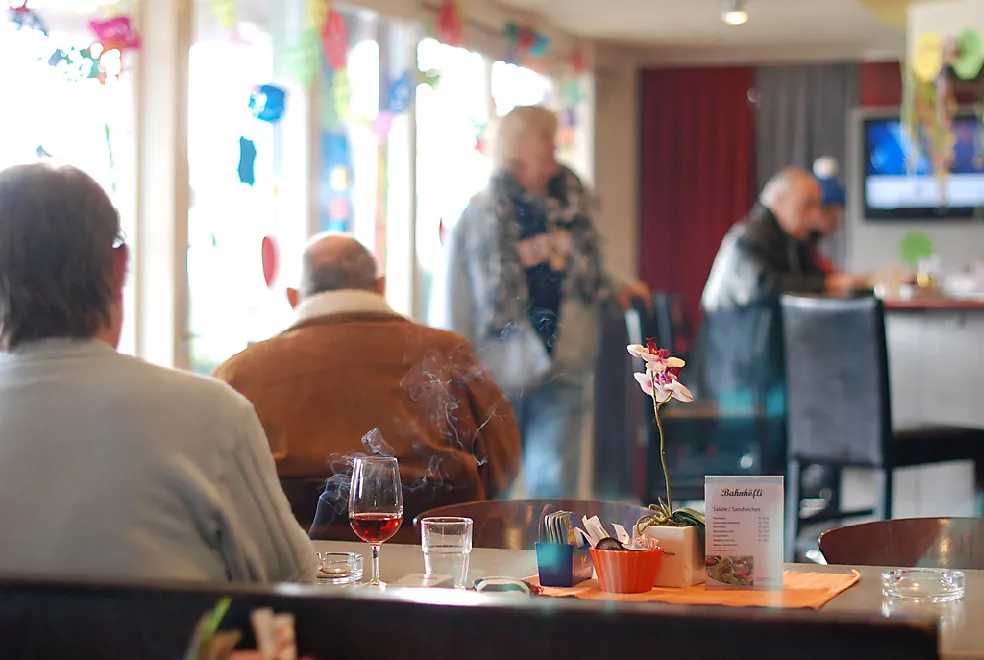 A person sits at a restaurant table that holds an ashtray with a burning cigarette in it while other patrons stand and sit nearby.