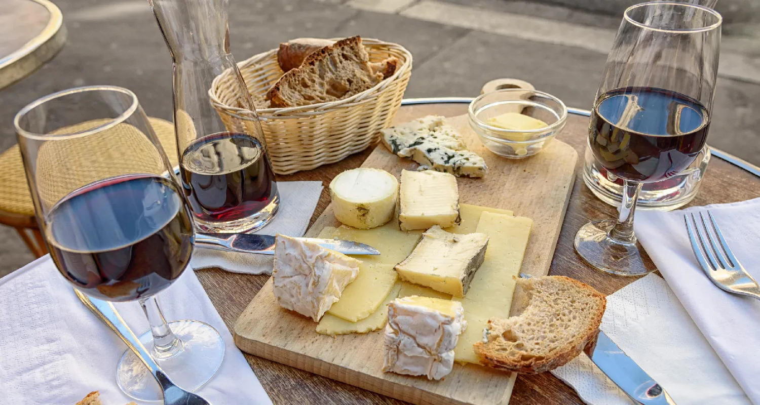 A carafe of wine and two glasses of wine on a table. There is also a basket of sliced bread and a board with different types of cheese on it.