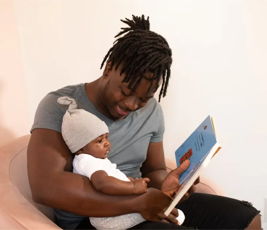 Photo of infant sitting in adult’s lap looking at book that the adult is reading to the infant.