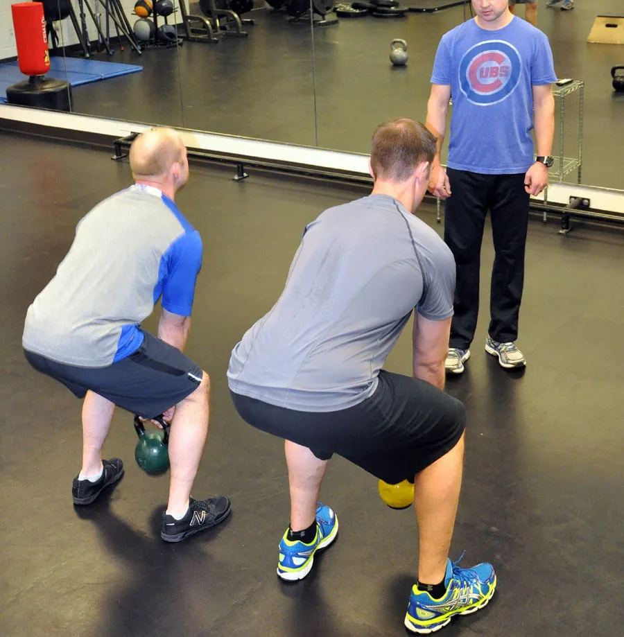 Two people perform kettlebell squats in a gym while a trainer observes.