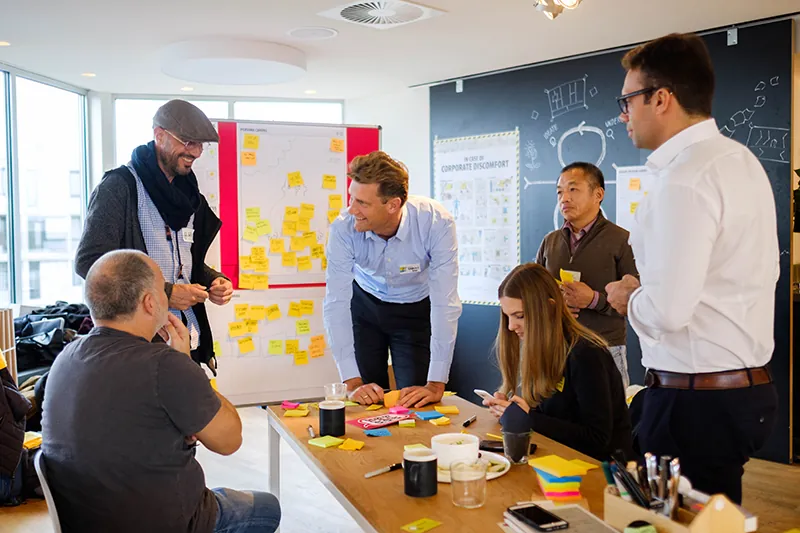 A group of people work around a table. They are talking and writing on stickie notes. Behind them is a flip board with stickie notes stuck to it and a chalkboard with writing and diagrams on it.