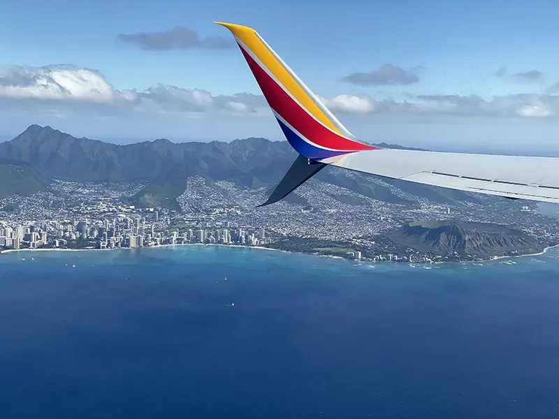 The wing of a Southwest Airline airplane flies over a body of water with a mountain in the distance. The plane is recognizable as Southwest due to the stripes from the logo on the wing.