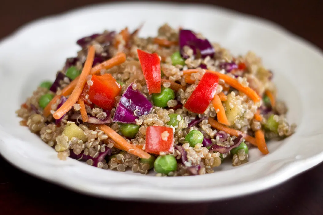 A bowl of cooked quinoa, diced purple cabbage, diced red bell peppers, peas, and julienned carrots, with a ginger soy dressing made with sesame oil, rice wine vinegar, green onion, and sesame seeds.