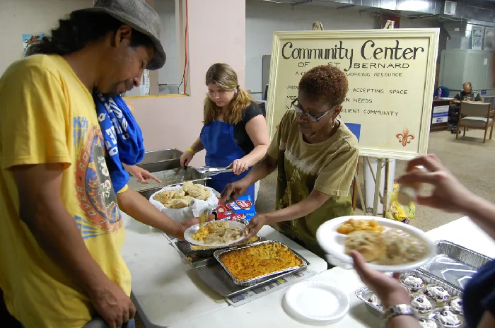 People scoop food from trays onto an individual’s plate. Behind them is a sign that says “Community Center of St. Bernard”.
