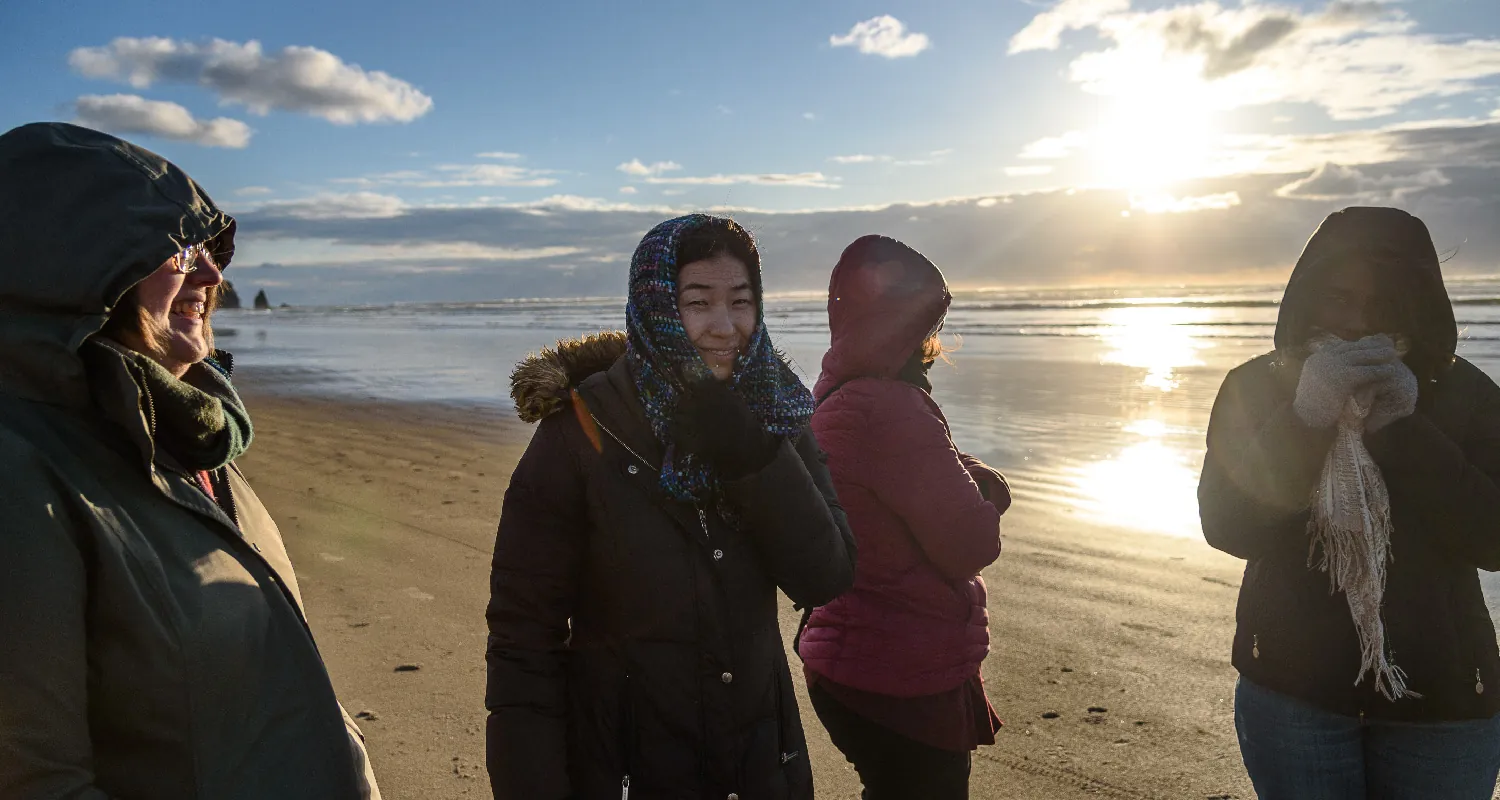 Four people wearing  scarves, gloves, and winter coats stand on the beach near the water. The two whose faces are visible are smiling.