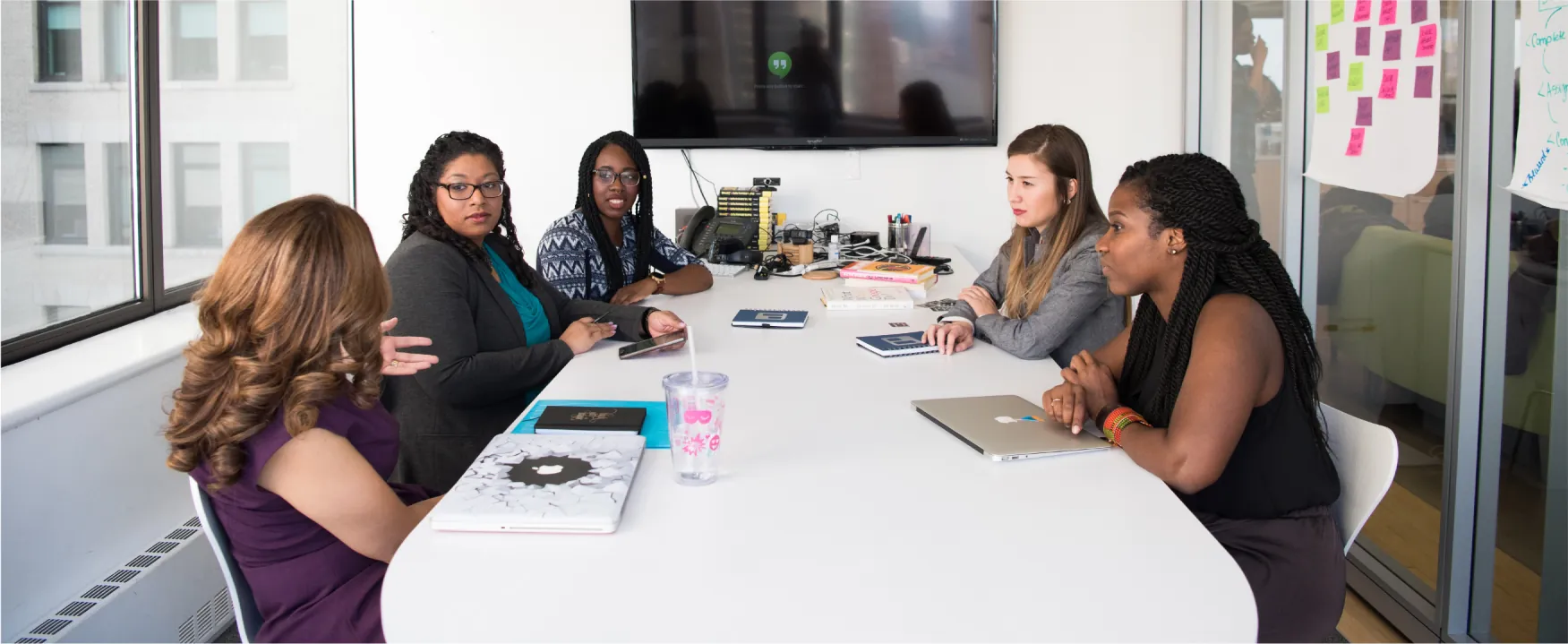 A group of women are gathered around a conference table with laptops and notebooks.