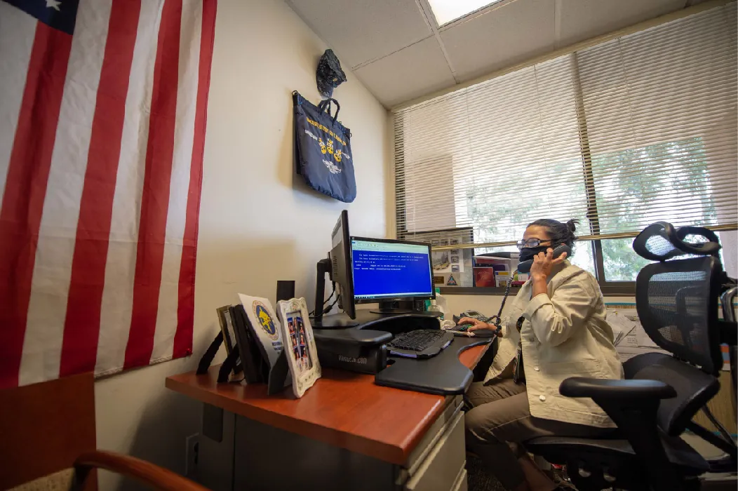 A person sits in a chair at a desk in front of a computer. They talk on the telephone while looking at the computer screen.