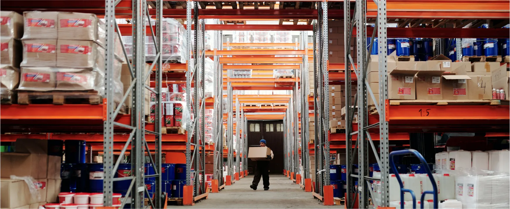 A person walks down the aisle of a warehouse holding a large box. Shelves on either side of the aisle are filled with various items.