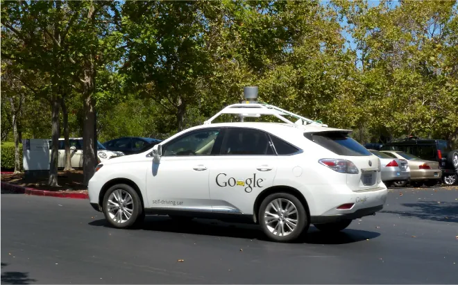 A car has “Google” written on the back and “self-driving car” written on the front door. A gray, round canister sits on a white framework on the top of the car.