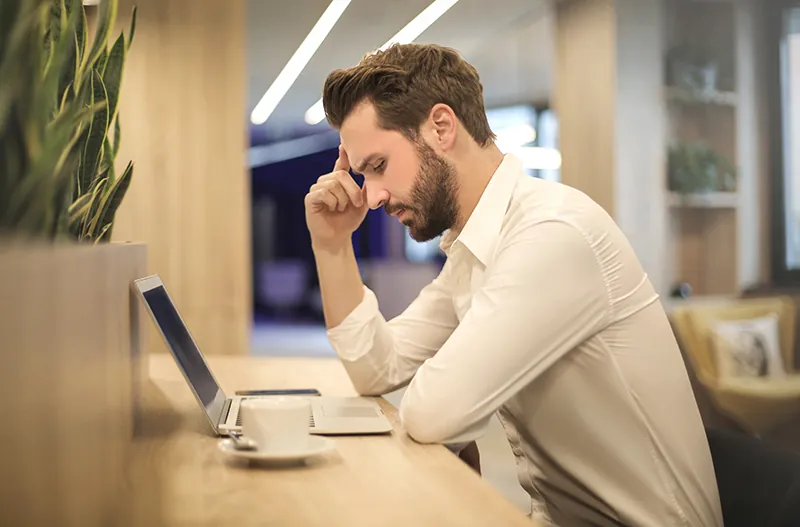 A photo shows the side view of a young man sitting at a table, looking stressfully at his laptop. A mobile phone and a coffee cup appear on either side of the laptop.