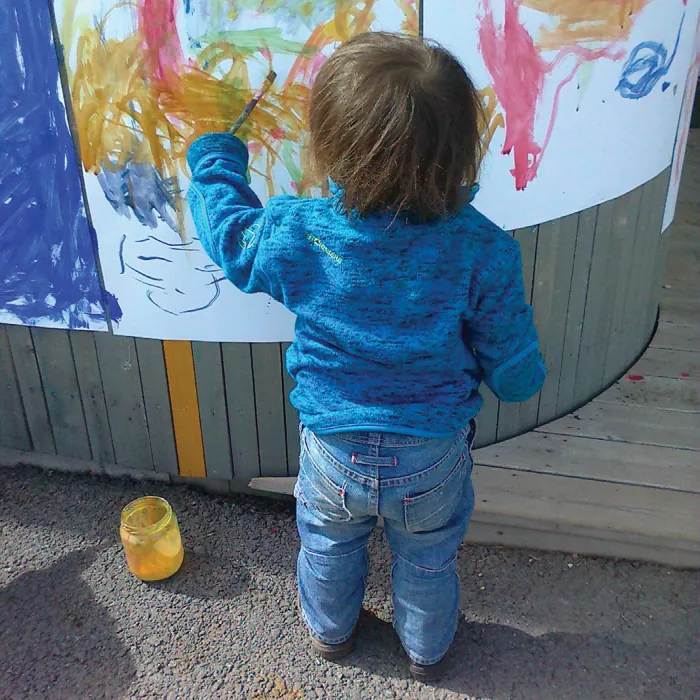 Photo of a young child painting on a white paper hanging on a fence.
