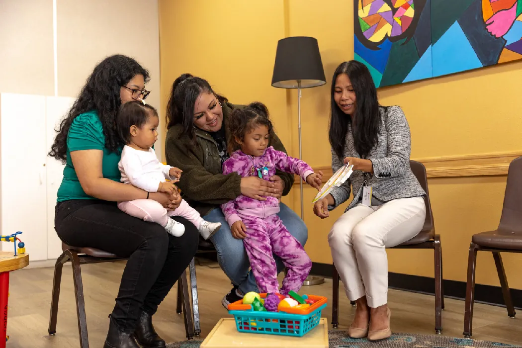 Two small children sit on the laps of two adults, who are seated in a large room. A third adult is seated next to them and holds up a paper. One of the children points toward something written on the paper.