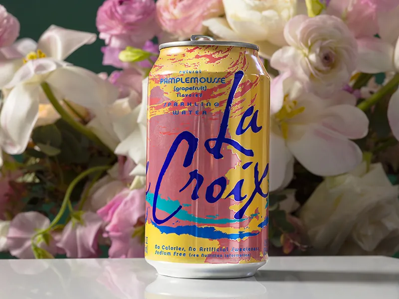A LaCroix can is in front of blooming white and pink flowers. The LaCroix can is bright yellow and pink with bright blue letters.