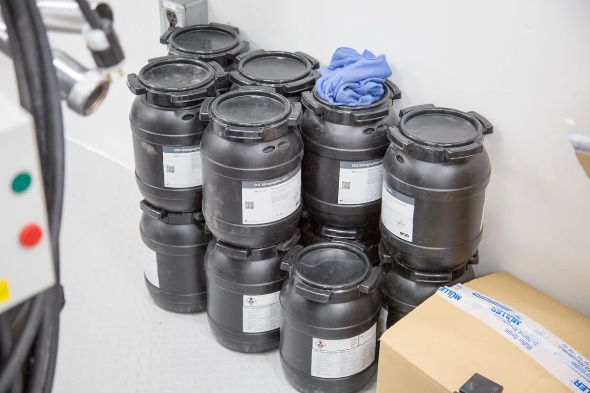 About 13 canisters with substantial sealed lids and detailed labels are stacked in two layers against a wall.
