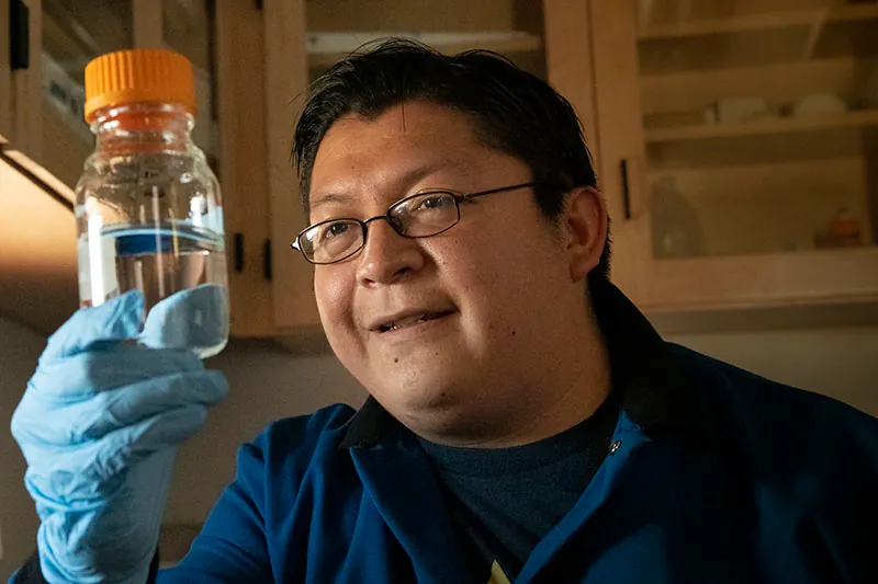 A photo shows a pharmaceutical expert wearing hand gloves examining a transparent solution inside a glass bottle held in his hand.