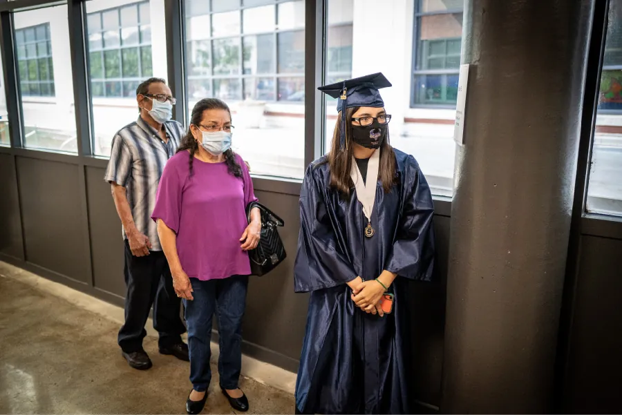 Three people stand together near windows. All of them wear face masks over their mouth and nose. One of the people is wearing a graduation cap and gown.