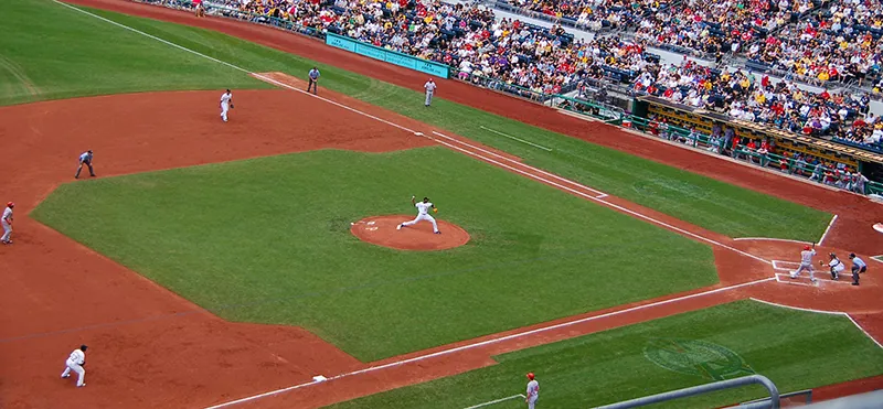 A baseball diamond is shown with a game in progress. The pitcher is pitching the ball to a batter at home plate.