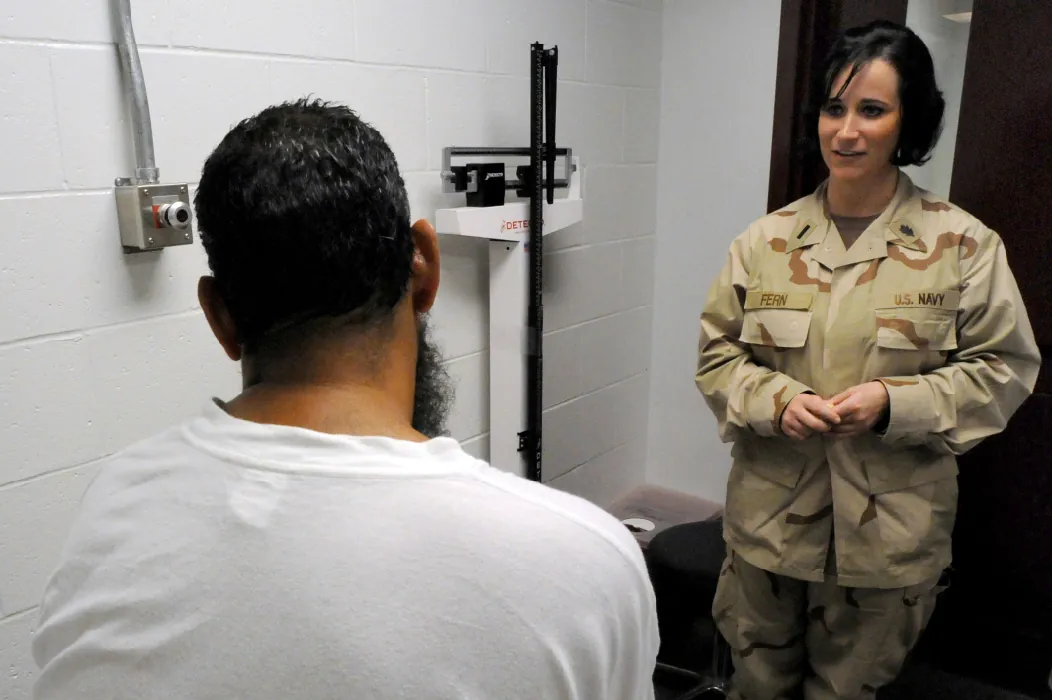 A nurse in navy fatigues stands across from a seated client in a room with a scale that measures height and weight.