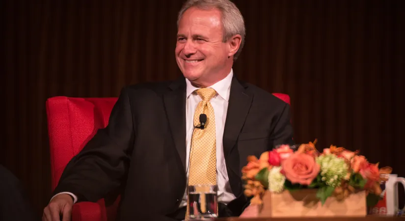 A photo of a smiling man with gray hair, with a microphone attached to his tie.