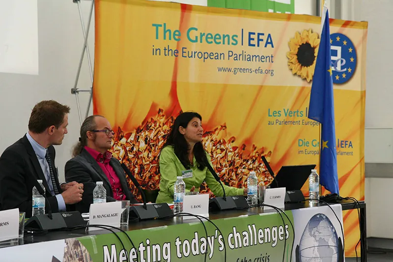 Three people sit behind a long table set with laptops, microphones, water bottles, and nametags. A sign hanging off the table reads “Meeting today’s challenges.” A large banner picturing a sunflower that hangs behind the people at the table reads “The Greens | EFA in the European Parliament,” and an EU flag stands next to the table.