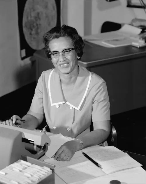 This black-and-white image shows Katherine Johnson smiling while sitting at a desk, working with a typewriter. She is wearing glasses and a dress with a white collar. Behind her, there is a filing cabinet and some papers on the desk, as well as what appears to be a map or chart on the wall.