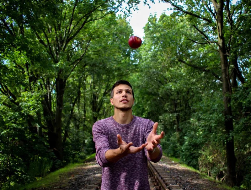 A young man in a purple sweater stands on railroad tracks in a forest, looking up as he juggles or catches a red apple above his hands. Lush green trees frame the scene.