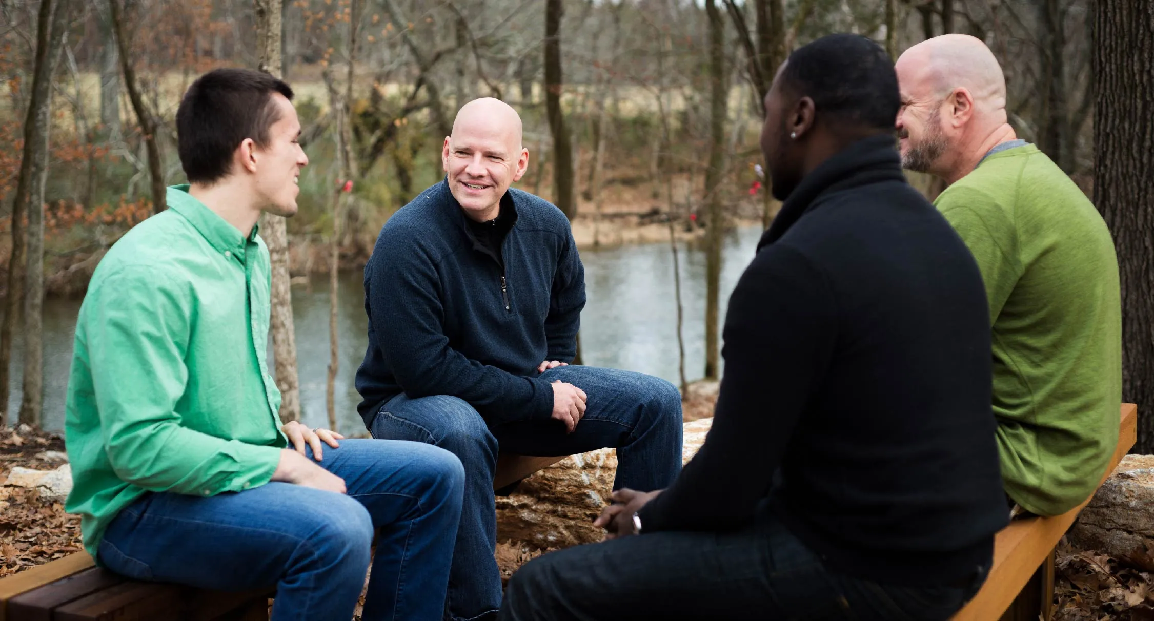 Four men sitting on a wooden bench in the forest by water talking.