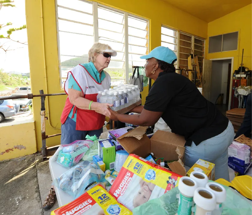 A community health worker stands behind a table stacked with supplies such as diapers, baby formula, and bottled water. They hand a case of bottled water to a person standing on the other side of the table.