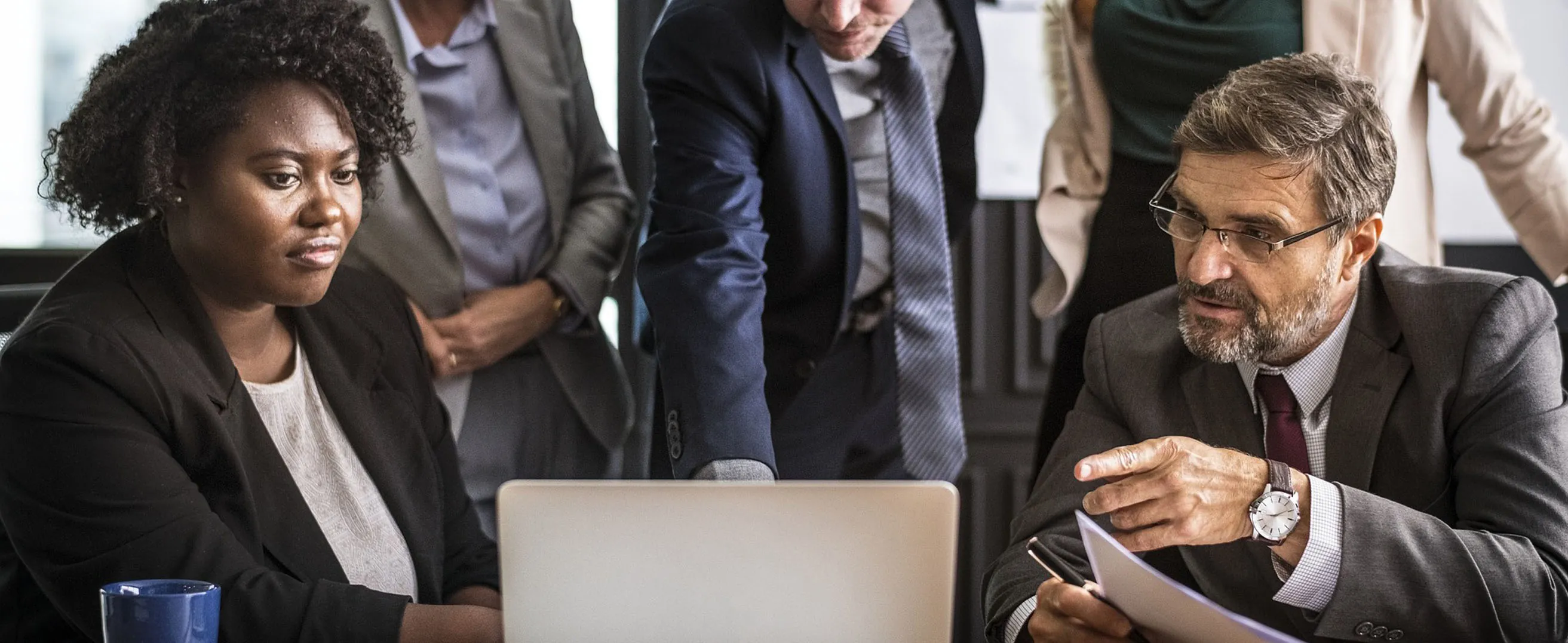 A photograph of  a group of men and women in business attire looking at a laptop screen.