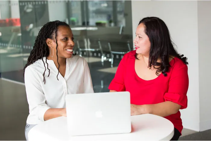 Two individuals converse at a table behind a laptop.