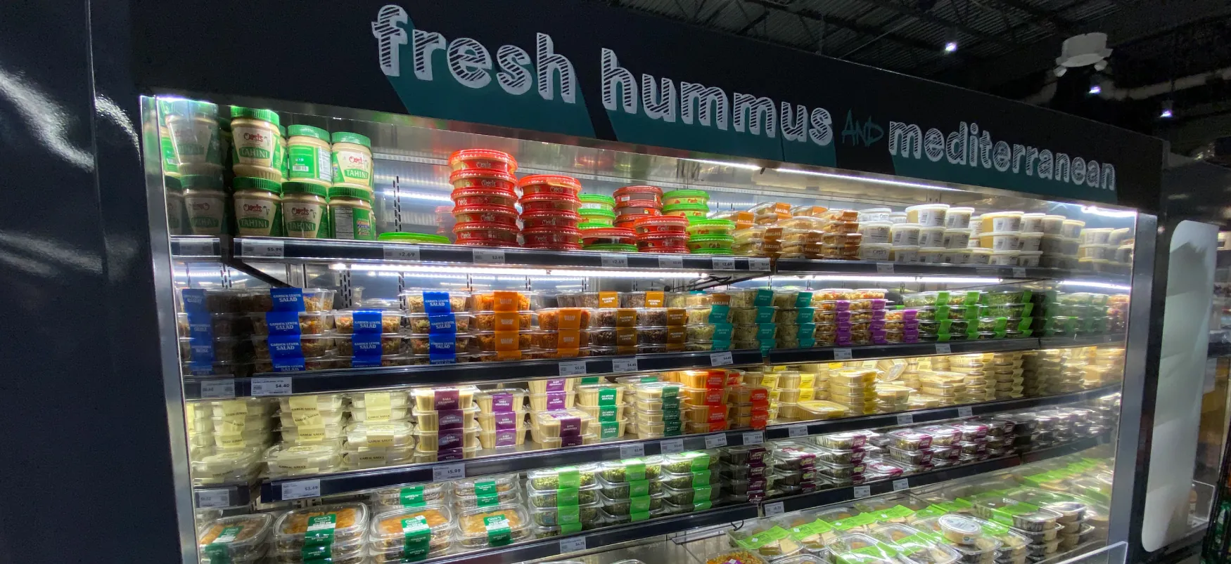 A large refrigerated section of a grocery store contains rows of small round containers of food. Above the rows are the words “fresh hummus and Mediterranean”