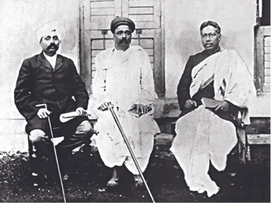 The photograph shows three men seated in chairs in front of a white house with shuttered doors. The man on the left wears a dark, long coat, pants, has a moustache, a white turban on his head and holds a cane in one hand a roll of papers in the other. The man in the middle wears a long white dhoti, has a moustache, a dark small cap, and holds a cane in one hand and a book in the other. The man on the right wears a dark coat and a large white cloth draped over his shoulder and covering his legs. He holds a book in his hand and wears glasses.