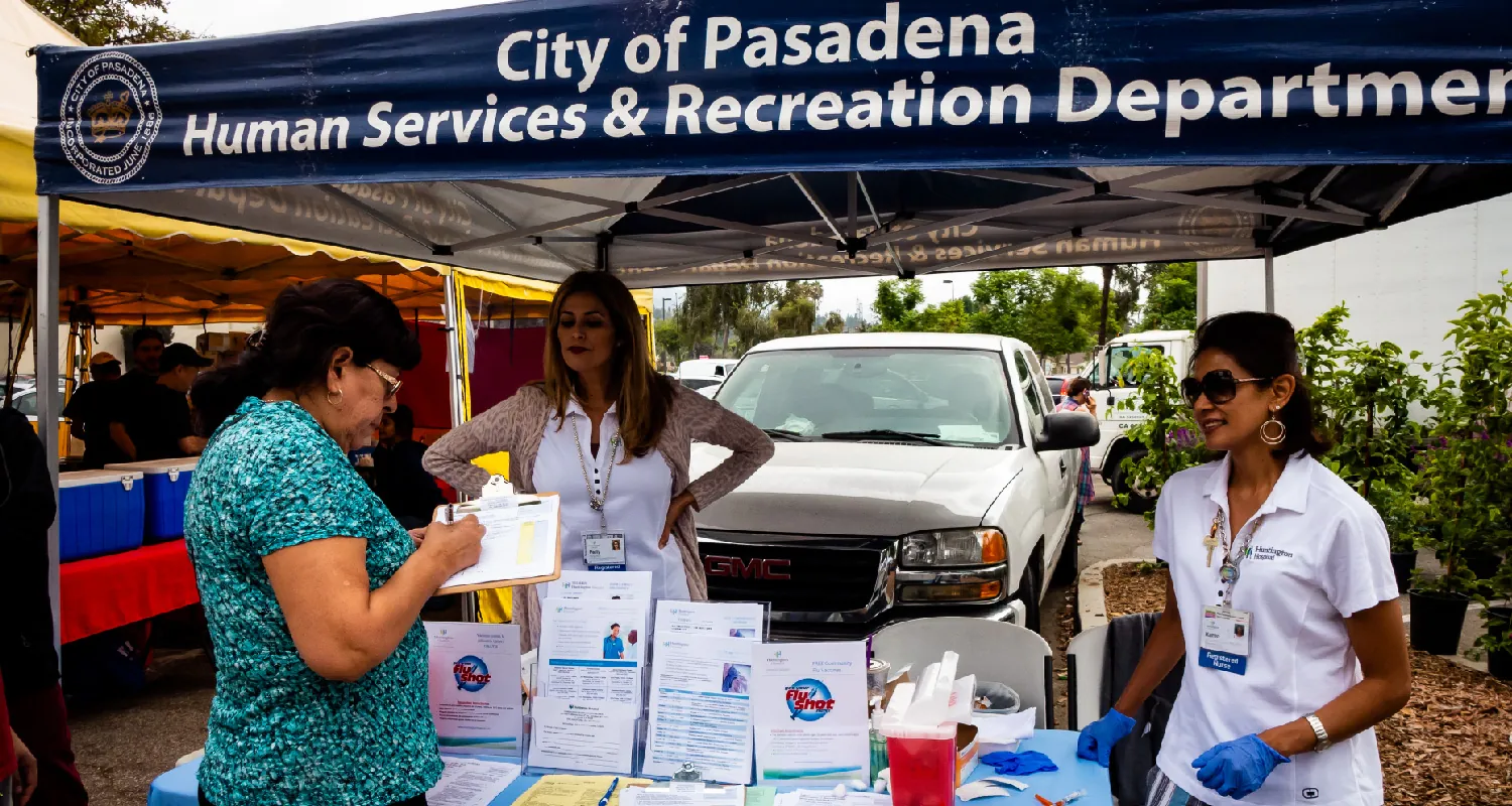 Two public health professionals stand near a table under an awning that says City of Pasadena Human Services and Recreation Department. The table has papers and medical supplies on it. A community member stands in front of the table filling out a document on a clipboard.