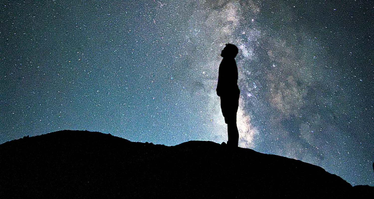 Photo of person standing on hill looking at night sky filled with stars and reflected clouds.