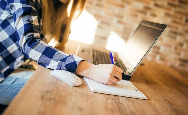 A student sits at a desk in front of a laptop computer. The student writes information on a notepad.