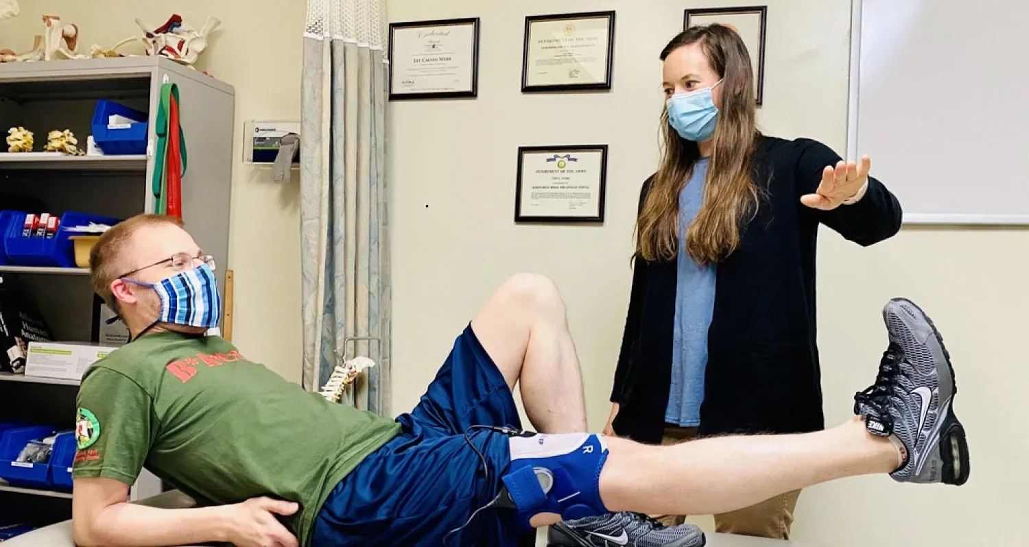 A physical therapist is working with a patient in a clinical setting. The patient, a young man wearing glasses, a green t-shirt, blue shorts, and a striped face mask, is lying on an examination table with one leg raised. The therapist, a woman wearing a blue face mask, blue shirt, and black cardigan, is standing beside the patient with her hand extended, guiding him through an exercise. The background features medical equipment, shelves with supplies, and framed certificates on the wall.