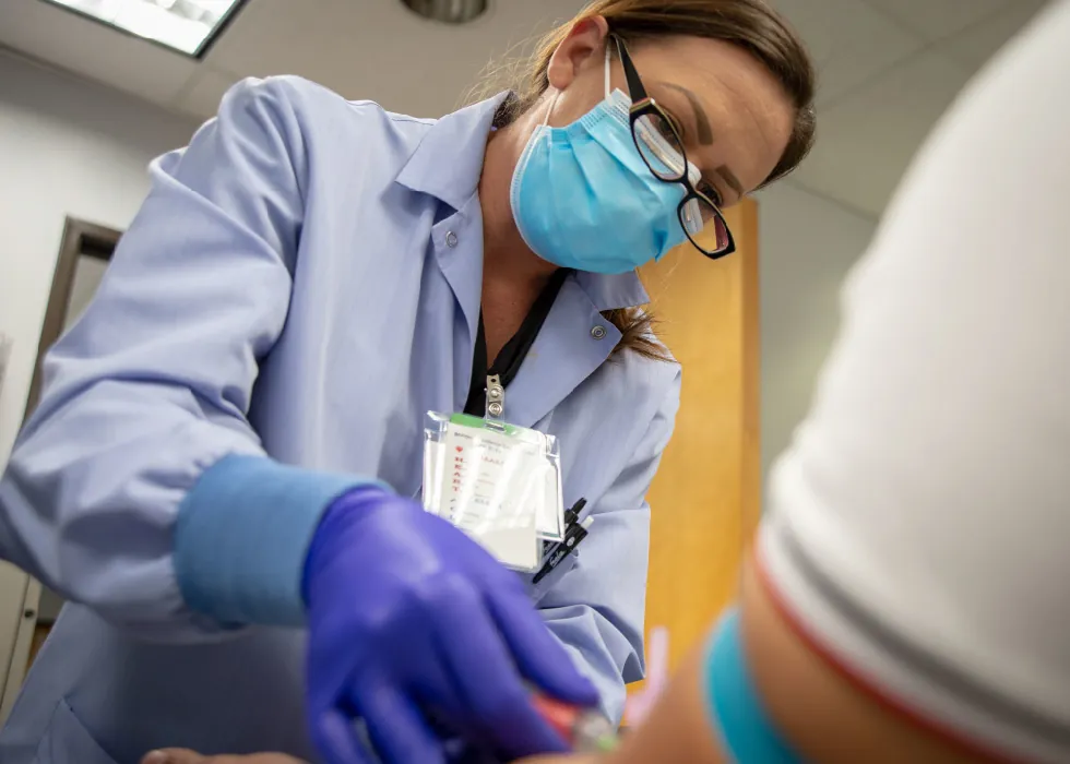 A medical professional wearing a lab coat, paper face mask, and blue gloves draws blood from a client’s arm.