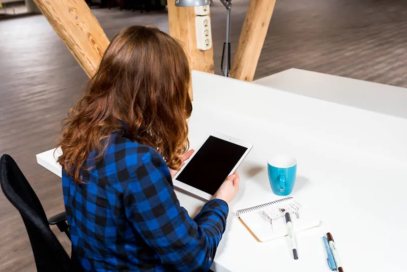 A seated student reads a tablet. A notebook, a coffee mug, and three pens reside on the desk.