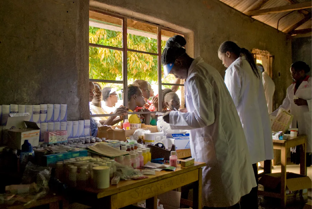 Medical personnel stand inside a building near a window. On a table near them are many different bottles of medicine. Children peer in the window.