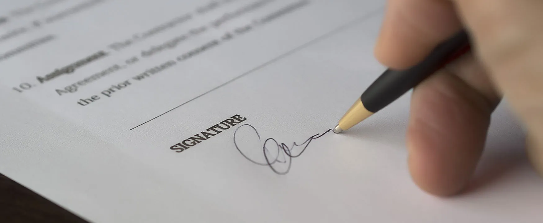 A close up of a signature line on a contract, with a person’s hand signing it.
