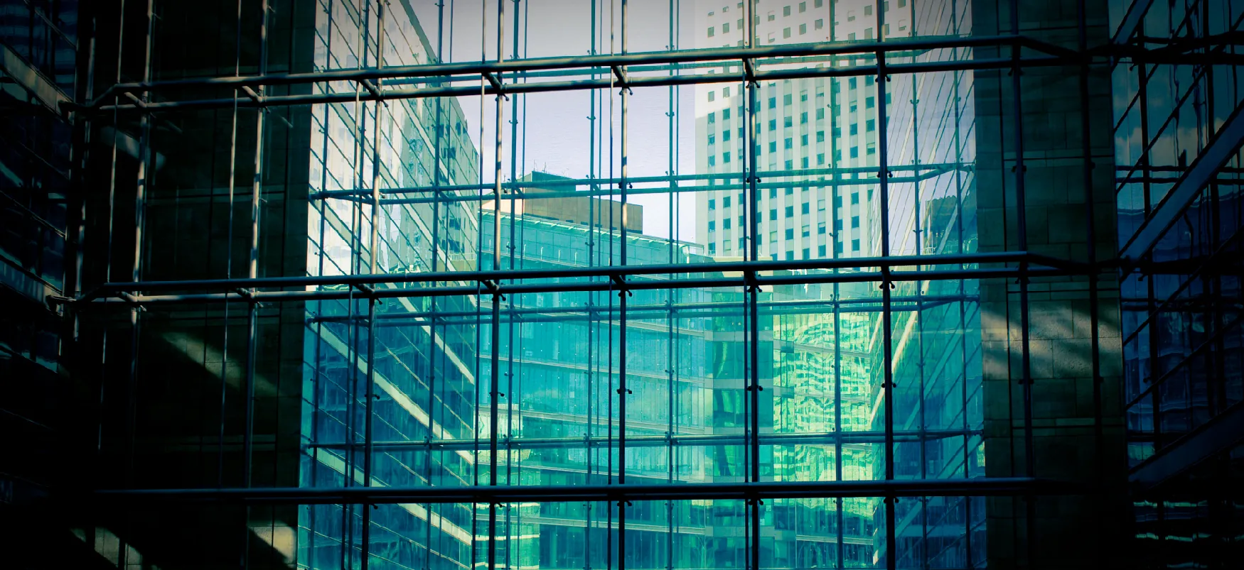 A building under construction has a reflective glass façade, which shows reflections of urban skyscrapers, and supporting metal rods in front.