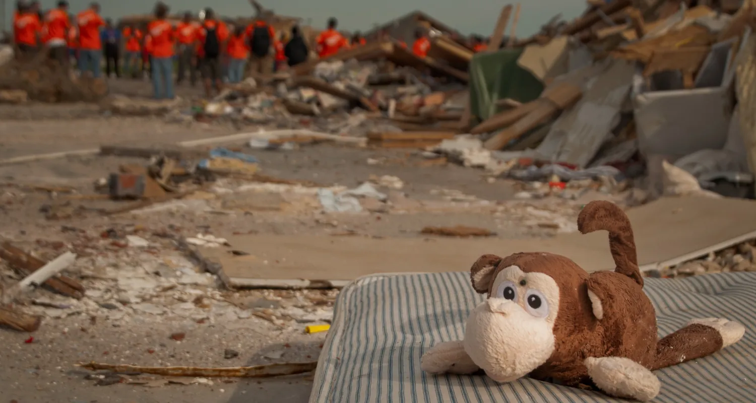 A color photograph shows a disaster area with cleanup workers in the background and a small child’s toy in the foreground.