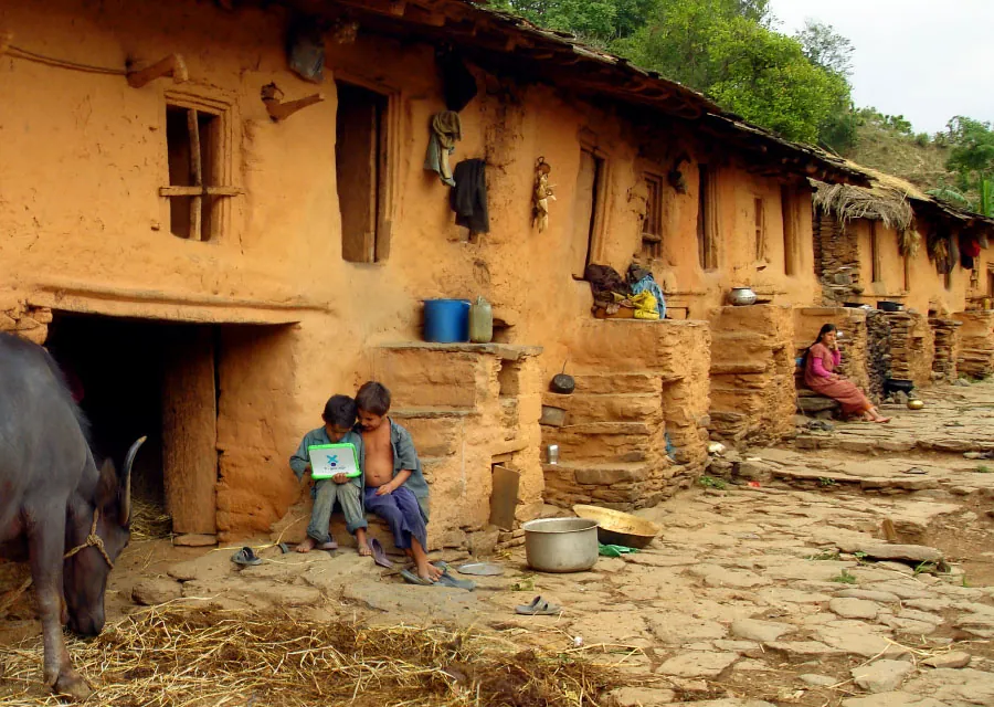 This image shows a rural setting with a row of mud houses in the background. Two children sit on the ground outside the house, one holding a laptop, and a buffalo is grazing nearby on the left. Various household items, such as pots and baskets, are scattered around the outdoor area. A woman sits farther down near the door of one of the houses.