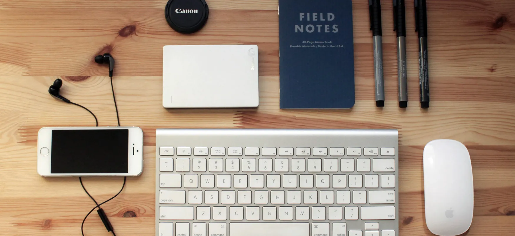 A notebook, three writing pen, a keyboard, a mouse, a mobile phone, lens cap of a digital camera, and a white colored small rectangular box, kept on a table.