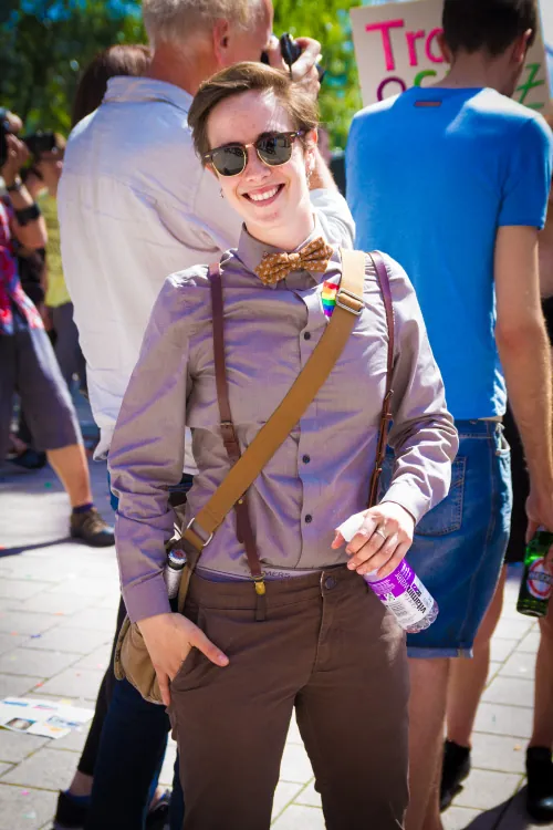 Photo of person with short hair, sunglasses, dressed in a purple, long sleeved shirt, brown pants, with a bow tie and suspenders, with no indication as to their gender.