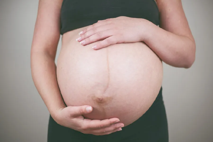 A pregnant woman cradles her abdomenal area with her hands. A narrow brown line is visible from the top of the abdomen to the belly button and then continues to the bottom of the abdomen.